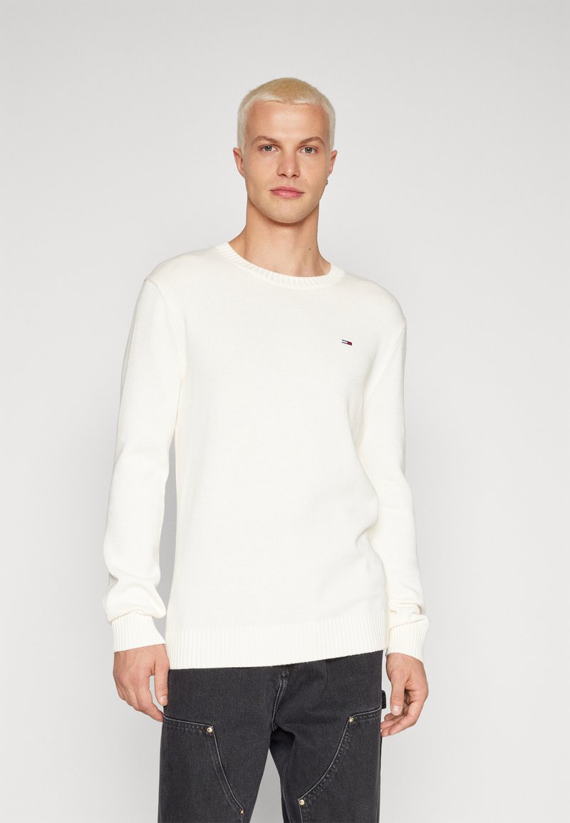Suéter blanco de punto con cuello redondo, puños y dobladillo acanalados, que presenta un pequeño logo en el pecho. El modelo lleva pantalones oscuros.