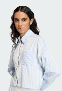 Pasekowa kurtka w jasnoniebieskim i białym kolorze. Posiada zamek błyskawiczny z przodu, kołnierz typu point oraz pojedynczą kieszeń na piersi z małym logo. Miękka tekstura tkaniny.