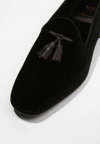 Mocassins en velours noir à la forme effilée, présentant une finition texturée et deux pampilles en cuir à l'avant pour les détails.