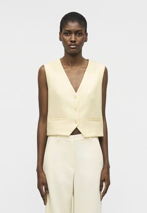 Donna che indossa un gilet senza maniche beige chiaro con bottoni e pantaloni abbinati a vita alta, su uno sfondo semplice.