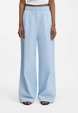 Person trägt hellblaue, weit geschnittene Hosen mit weißen vertikalen Streifen, kombiniert mit schwarzen Sandalen mit offenem Zehenbereich und einem weißen, kurz geschnittenen Oberteil.