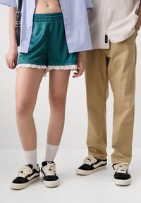 Teal satin shorts med blondekant, brukt med en lys blå oversized skjorte med knapper. Lyse beige bukser ved siden av, begge med svarte sneakers.