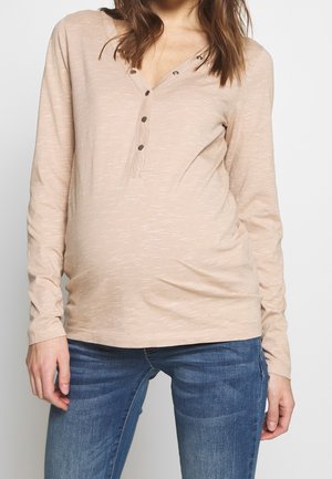 Zwangere vrouw die een beige langmouwen henley-shirt en blauwe spijkerbroek draagt, staand tegen een effen lichte achtergrond.