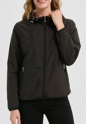 Femme portant une veste à capuche noire zippée avec des poches latérales zippées, tenant le col près de son menton, souriante.