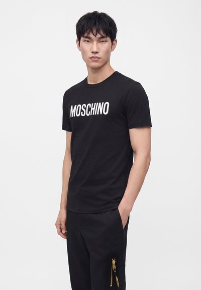 Νέος άντρας που φοράει μαύρο t-shirt Moschino και μαύρο παντελόνι με χρυσή λεπτομέρεια φερμουάρ, στέκεται μπροστά σε ένα απλό λευκό φόντο.