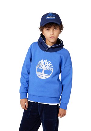 Sweatshirt bleu avec un logo blanc et capuche, associé à un pantalon en velours côtelé bleu marine. Le sweatshirt a une coupe décontractée et des poignets côtelés.