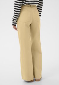 Pantalon large jaune en coton, avec taille haute, deux poches arrière et texture lisse, porté avec un haut à rayures à manches longues.