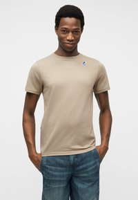 Άνδρας με σκούρο δέρμα που φοράει μπεζ κοντομάνικη μπλούζα με λογότυπο στο στήθος και μπλε τζιν, στέκεται με τα χέρια στις τσέπες μπροστά σε λευκό φόντο.