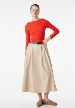 Kvinde iført en klar orange langærmet top, beige højtaljet midi nederdel med sort bælte og brune loafers, stående med den ene hånd på hoften.