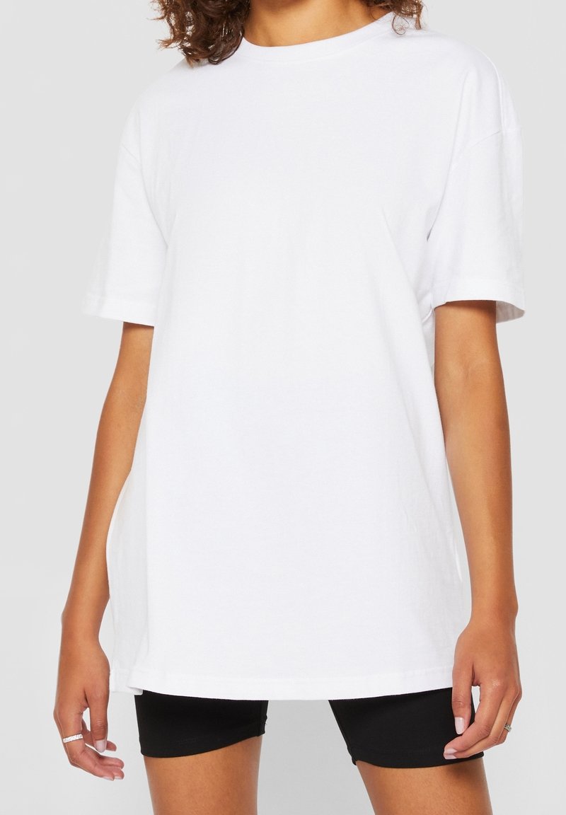 Femme portant un t-shirt blanc ample à manches courtes et un short noir ajusté, debout devant un fond clair uni.