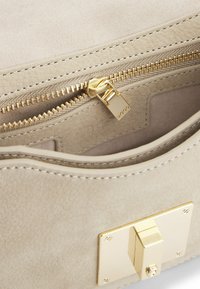 Beige mockasintö bag inredning med en guldkedja och detaljer. Har en slät textur, rundade kanter och ett litet spänne för stängning.