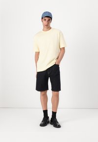 Short-sleeve light yellow t-shirt, dark denim shorts, black loafers, and a blue cap. Model stands with hands in pockets against a white background.