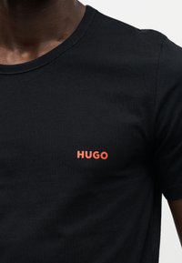 Close-up af en person iført en sort t-shirt med ordet "HUGO" trykt med små orange bogstaver på brystet.