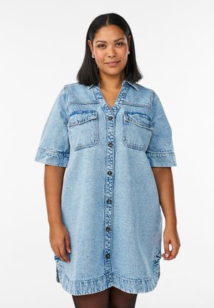 Kvinde med skulderlangt mørkt hår iført en lyseblå denimkjole med korte ærmer og knapper foran samt forlommer.