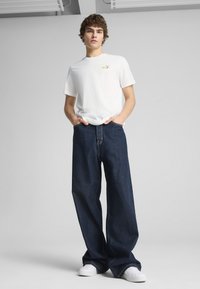 Camiseta blanca con un pequeño logotipo de color, combinada con unos vaqueros de pierna ancha en azul oscuro. El modelo lleva zapatillas blancas, de pie contra un fondo claro.