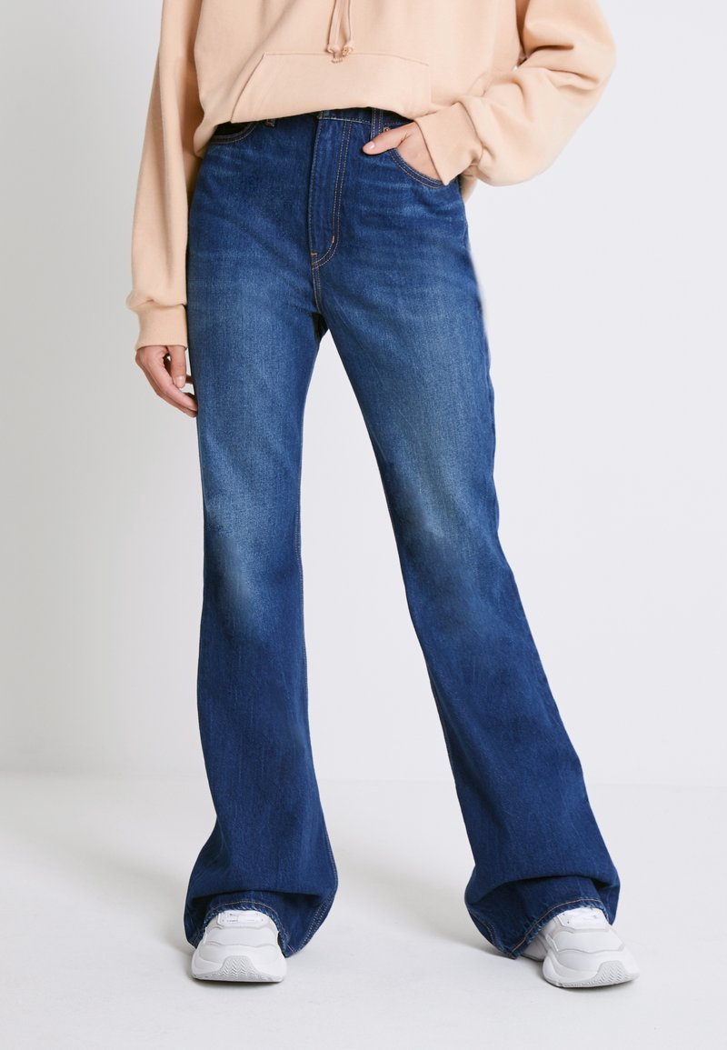 Personne portant un jean flare taille haute bleu foncé, un hoodie beige et des baskets blanches, debout avec une main dans la poche.