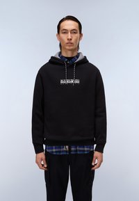 Felpa nera di cotone con fodera grigia, con un logo bianco centrato. Indossata sopra una camicia a quadri blu, abbinata a pantaloni cargo scuri.