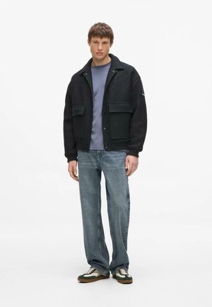 Noor mees seisab, seljas must jakk suurte esitaskutega, hall särk, laiad sinised teksad ja rohelised-valged tossud.