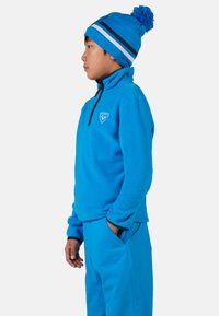 Giacca di pile blu con colletto alto e logo sul petto, abbinata a pantaloni blu coordinati e un berretto blu con strisce bianche e pompon.