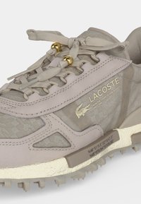 Сникърс в цвят топъл сиво-кафяв на Lacoste с медни капси, текстурирана тъкан, лого и текст, както и координати, отпечатани на бежовата подметка близо до петата.