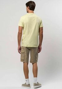 Férfi világos sárga rövid ujjú pólóban, bézs cargo nadrágban, fehér zokniban és bézs sportcipőben áll, háttal nézve egy sima háttér előtt.