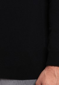 Czarny dziany sweter z prążkowanym dołem i mankietami, pokazany na szarym materiale, podkreślający jego fakturę i długie rękawy.