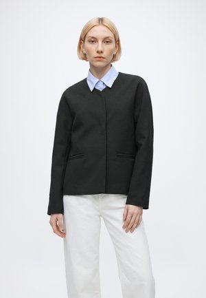 Femme aux cheveux blonds courts portant une veste noire par-dessus une chemise bleu clair à col et un pantalon blanc, debout devant un fond blanc uni.