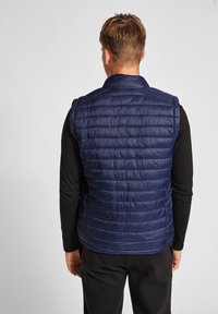 Marinblå puffväst med horisontell sömnad, hög krage och ärmlös design. Lättviktig, slät textur, del av en avslappnad outfit.