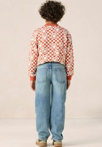 Enfant portant un sweat-shirt rouge et blanc à motifs de visages souriants, associé à un jean bleu ample et des baskets beiges.