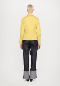Veste ajustée jaune avec des manches longues et des accents de boutons, associée à un pantalon flare en denim foncé muni de poignets gris.