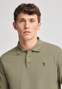 Khakifärgad polo med korta ärmar, treknappsöppning och broderad logotyp på bröstet. Tillverkad av bomull med en slät textur.