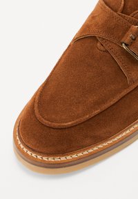 Smeđe suède slip-on cipele s okruglim vrhom, šivenim detaljima i kontrastnom gumiranom potplatom. Dizajn uključuje pojas s ukrštenim detaljima.