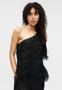 Μαύρο τοπ με έναν ώμο και διακοσμητικές φούντες. Μαλακή υφή, άνετη εφαρμογή και μινιμαλιστικός σχεδιασμός. Διακριτική λάμψη που ενισχύει την οπτική εμφάνιση.