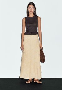 Jupe beige à texture ondulée, assortie à un débardeur noir ajusté. Les accessoires comprennent un sac en perles marron et des sandales plates marron.