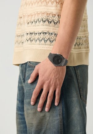 Montre noire avec un cadran rond, bracelet en métal argenté et grands marqueurs d'heure, portée sur une main reposant sur un jean en denim bleu. Le haut présente un design en tricot beige.