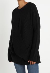 Pull en maille côtelée noir avec une coupe ample, épaules tombantes et manches longues. Possède une texture douce et un drapé subtil à l'ourlet.