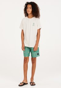 Lichtbeige t-shirt met groen logo, gecombineerd met groene shorts met een kleine print. Zwarte slippers maken de outfit compleet.