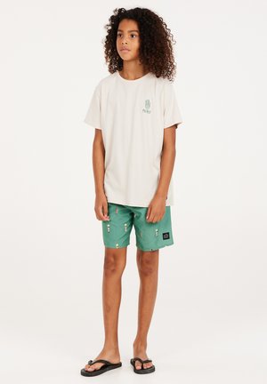 Světle béžové tričko s zeleným logem, doplněné zelenými šortkami s malým potiskem. Černé žabky dotvářejí celý outfit.