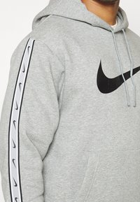Gry hooded sweatshirt i en bomullsmix, med en svart Nike-logotyp och svart-vitt märkesband längs ärmarna. Framlommer synlig.