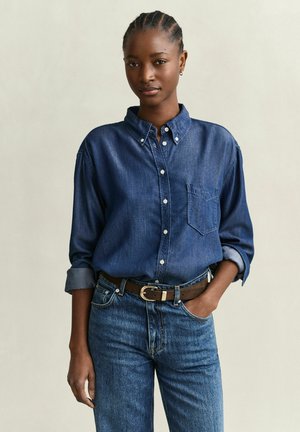 Junge Frau mit geflochtenem Haar, die ein Jeanshemd trägt, in hoch taillierte Jeans mit braunem Gürtel gesteckt, vor einem schlichten Hintergrund stehend.