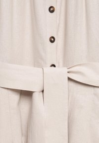 Beige mekko, jossa on napitettava etuosa, ruskeat napit ja yhteensopiva vyötärönauha. Valmistettu teksturoidusta kankaasta, jossa on sileä pinta.