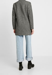 Cappotto lungo grigio in misto lana con un design su misura, abbinato a jeans a gamba larga azzurri e stivaletti neri. Dettagli con maniche arrotolate.