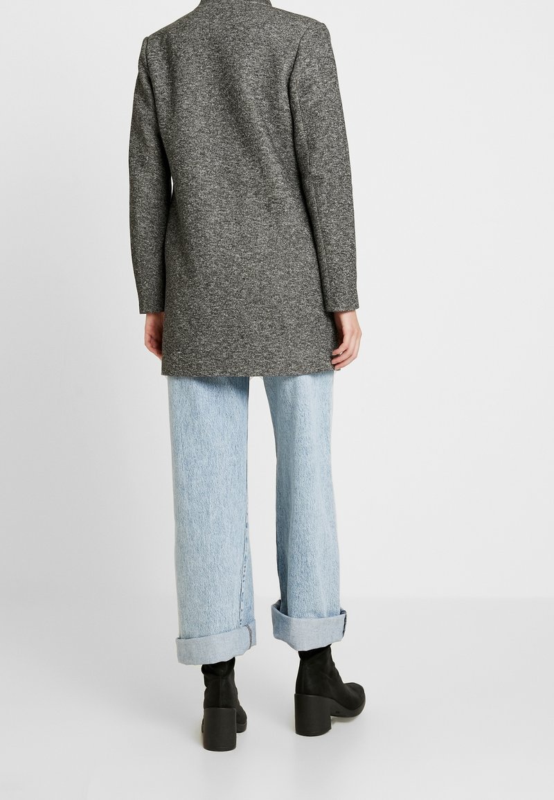 Cappotto lungo grigio in misto lana con un design su misura, abbinato a jeans a gamba larga azzurri e stivaletti neri. Dettagli con maniche arrotolate.