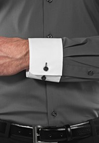 Camicia dress grigia con polsini bianchi; bottoni neri sui polsini e sulla pattina; tessuto liscio; cintura in pelle con fibbia lucida.