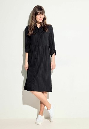 Schwarzes, knopfbares Kleid mit vertikalen Streifen, hochgekrempelten langen Ärmeln und taillierter Taille. Kombiniert mit weißen Sneakers.