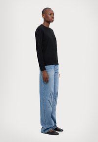 Maglione nero in maglia, jeans a gamba larga azzurri con motivo a logo e scarpe slip-on nere. Texture liscia con una vestibilità rilassata.