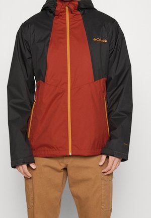 Homme portant une veste Columbia rouge et noire avec des accents de fermeture éclair jaunes et un pantalon cargo beige, mains détendues le long du corps.