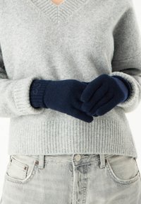 Navy Wollhandschuhe mit gerippten Bündchen, getragen von einer Person in einem hellgrauen V-Ausschnitt-Pullover, der flache Nähte und eine weiche Textur zeigt.