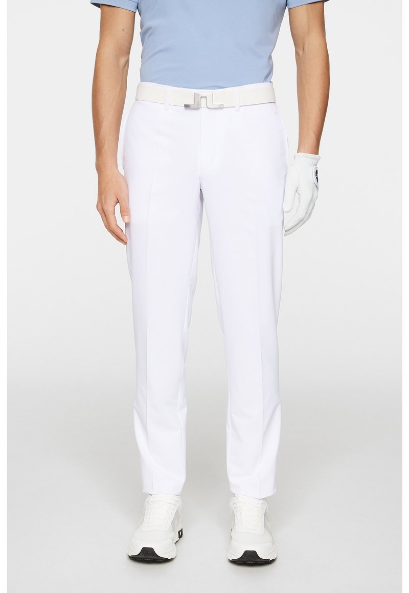 Białe spodnie golfowe o eleganckim kroju i dopasowanym fasonie, w połączeniu z jasnoniebieską koszulą i białym paskiem. Na prawej ręce założone są teksturowane białe rękawiczki.
