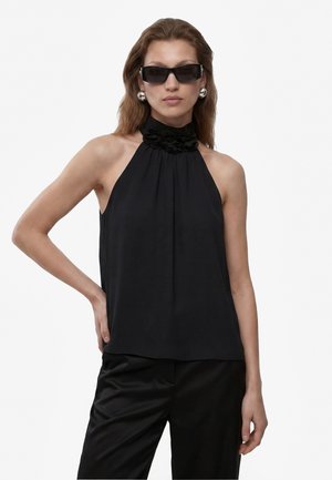 Femme portant un haut noir sans manches à col fleuri, un pantalon noir, de grandes boucles d'oreilles argentées et des lunettes de soleil noires rectangulaires sur fond blanc.